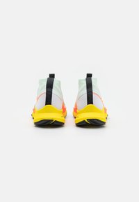 Ein Paar Sportschuhe mit gelben Sohlen, weißen und orangefarbenen Oberteilen sowie schwarzen Zuglaschen, von hinten auf einem weißen Hintergrund betrachtet.