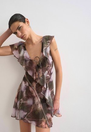 Frau mit dunklen Haaren in einem gebundenen, gerafften, lila-grünen, blumigen Minikleid, die vor einer schlichten weißen Wand steht und einen Arm hinter dem Kopf hebt.