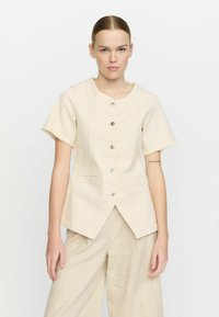 Beige kortärmad jacka med rund halsringning, sex knappar framtill, två sidofickor och rå kantdetaljering på ärmarna.