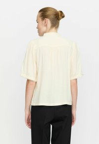 Helles Cream-farbenes Blusenshirt mit kurzen Ärmeln, Kragen, strukturiertem Stoff und zarter Spitze an den Ärmeln, kombiniert mit schwarzen Hosen.