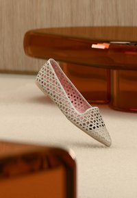 Γυναικείο μπεζ επίπεδο παπούτσι με γεωμετρικό σχέδιο κοπής και ροζ εσωτερικό, τοποθετημένο μπροστά από πορτοκαλί διαφανή έπιπλα σε μπεζ χαλί.