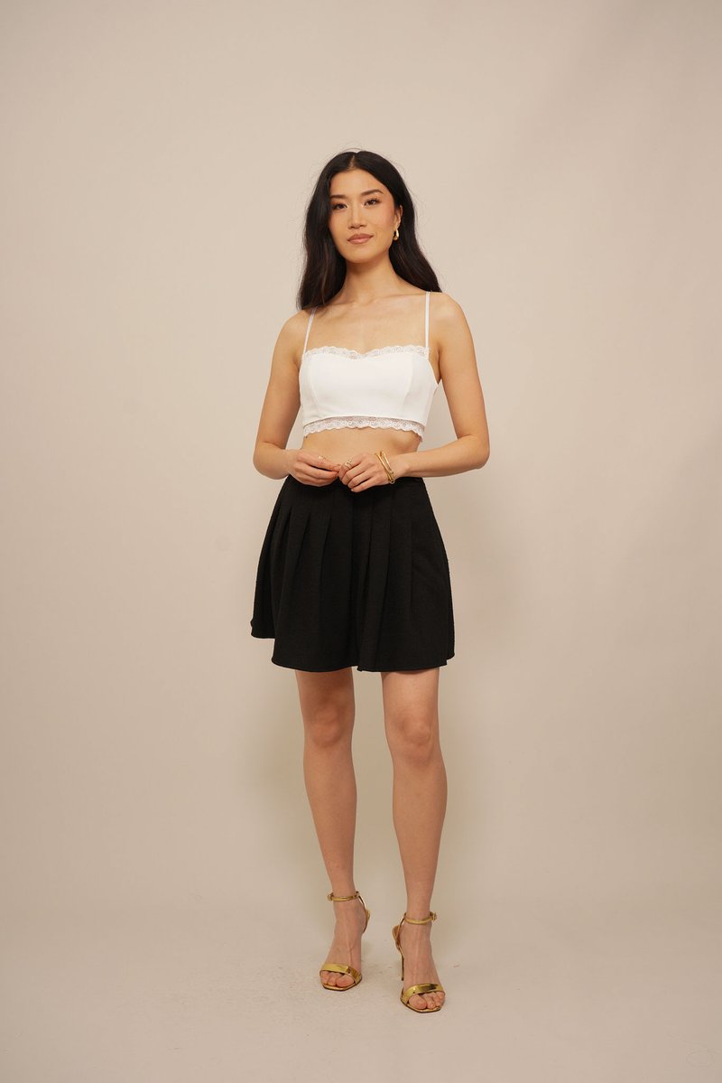 Crop top bianco con bordo in pizzo abbinato a una mini gonna plissettata nera. Modella che indossa sandali con tacco dorati, in piedi su uno sfondo neutro.