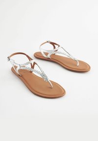 Sandali piatti con suola marrone e cinturini intrecciati metallici argento. Presentano un design con listino per le dita e una cinghia alla caviglia regolabile.