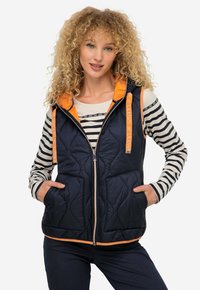 Gilet trapuntato blu navy con dettagli arancioni e cappuccio. Presenta una chiusura frontale con zip, due tasche e un orlo a contrasto. Indossato sopra una maglietta a righe a maniche lunghe.
