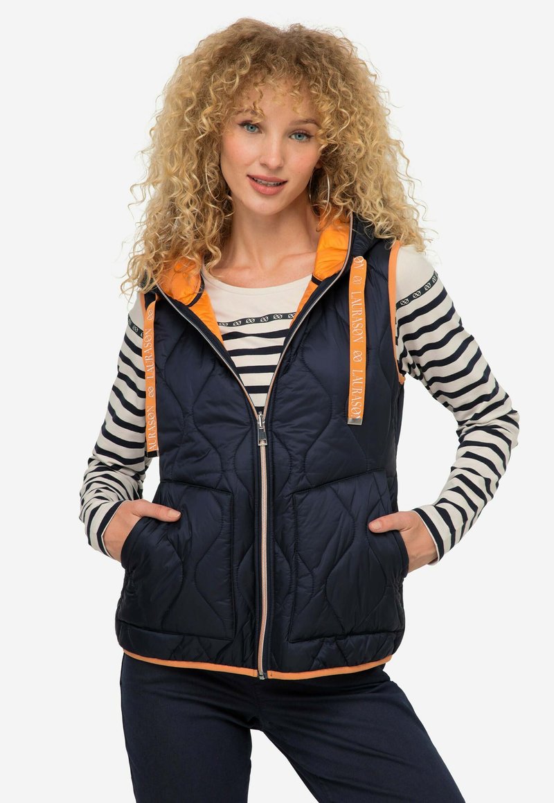 Gilet trapuntato blu navy con dettagli arancioni e cappuccio. Presenta una chiusura frontale con zip, due tasche e un orlo a contrasto. Indossato sopra una maglietta a righe a maniche lunghe.