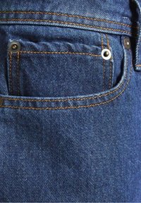 Vaqueros de mezclilla en azul oscuro, con costuras naranjas en contraste. Presentan un bolsillo delantero con acentos de remaches metálicos y un corte limpio.