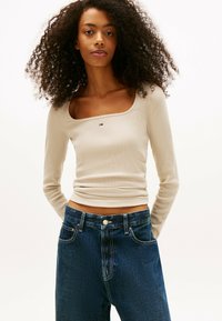 Joven mujer con cabello rizado, vistiendo una camiseta de manga larga de canalé beige y pantalones vaqueros de talle alto azul oscuro, de pie con las manos detrás de la espalda.