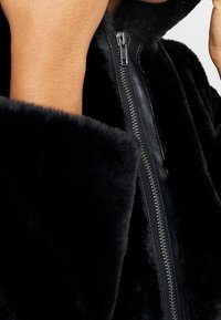 Czarna kurtka z futrzanym futerkiem ze sztucznego futra, z zamkiem błyskawicznym z przodu, kapturem i teksturowanymi rękawami. Projekt łączy miękkie futro i gładki skórzany zamek.