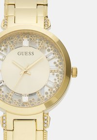 Zlatý GUESS hodinky s okrúhlym puzdrom, textúrovaným ciferníkom s kryštálovými akcentmi a náramkom s odkazmi s ďalšími trblietavými detailmi.