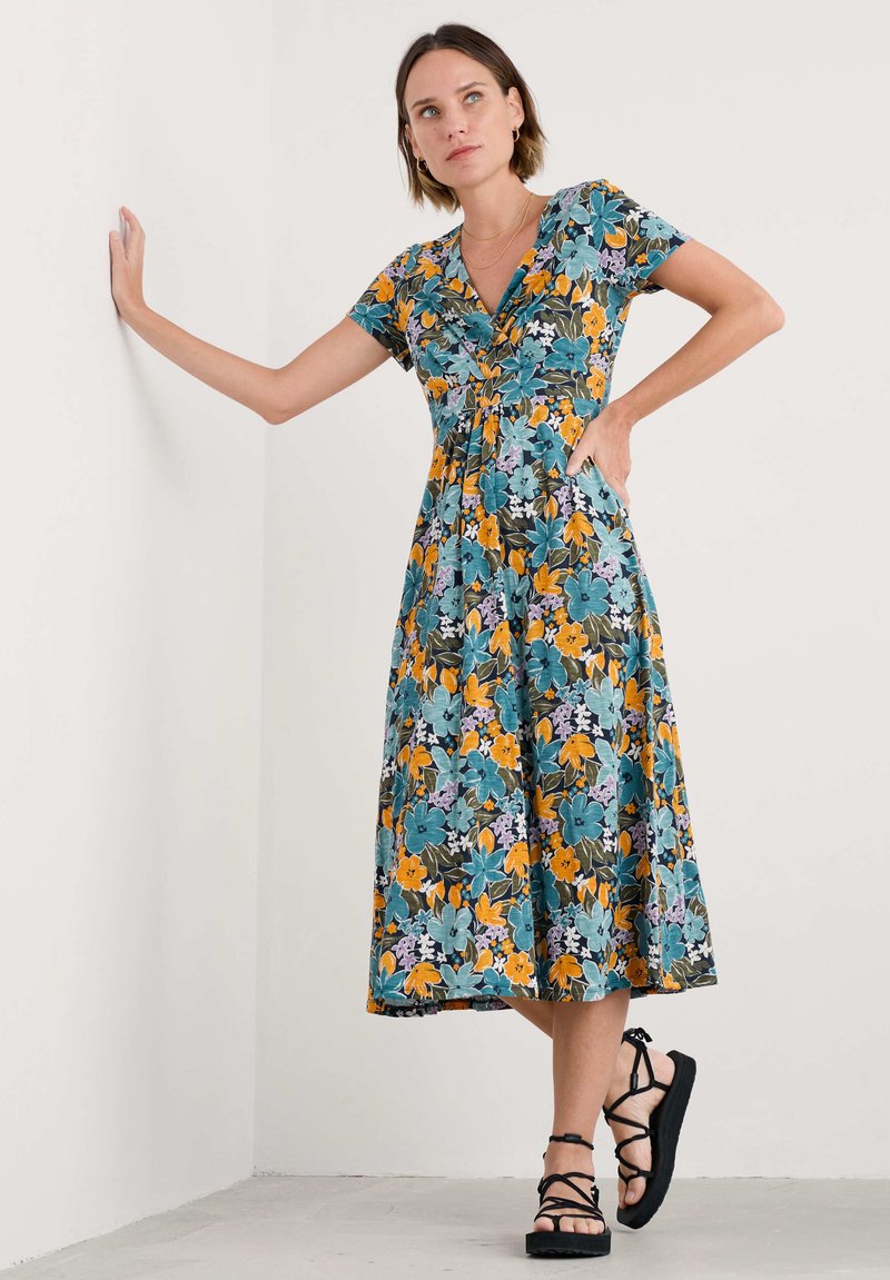 Blumenkleid in Midi-Länge mit einem Türkis-, Schwarz- und Orangemuster, kurzen Ärmeln, V-Ausschnitt und einem fließenden Saum. Tragen mit schwarzen Riemchensandalen.