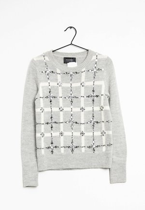 Pull en maille gris clair avec motif à carreaux blancs et perles argentées décoratives, présenté sur un cintre noir sur fond blanc.