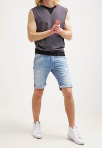 Sudadera sin mangas gris con un cuello negro, combinada con pantalones cortos de mezclilla claros y desgastados. Zapatillas blancas completan el atuendo.