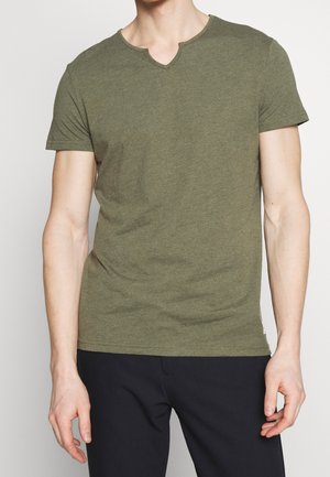 Mand iført olivengrøn kortærmet V-hals T-shirt og sorte bukser, stående mod en ensfarvet baggrund.