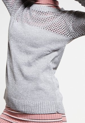 Femme portant un pull en maille gris clair avec un empiècement ajouré et des manches, par-dessus un haut rose côtelé et une jupe rayée, posant avec les bras levés.