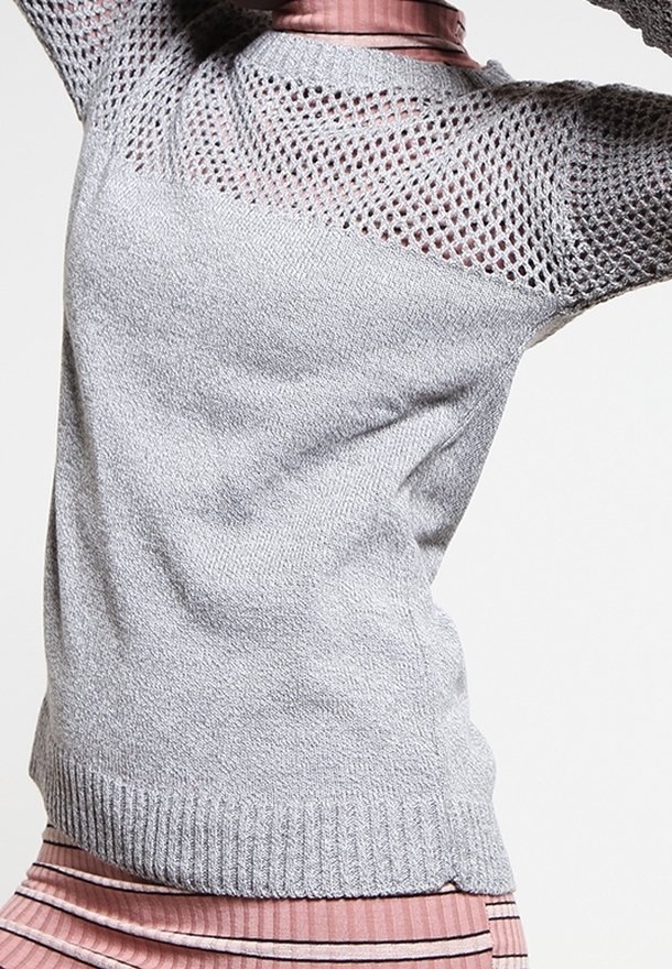 Femme portant un pull en maille gris clair avec un empiècement ajouré et des manches, par-dessus un haut rose côtelé et une jupe rayée, posant avec les bras levés.
