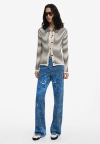 Gestreifter Strickcardigan in Creme und Marineblau, mit Kragen und Knopfschluss. Blaue gemusterte weit geschnittene Jeans und schwarz-weiße Schuhe runden den Look ab.