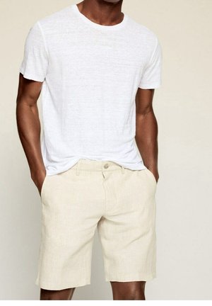 Mann trägt ein schlichtes weißes Kurzarm-T-Shirt und beige knielange Shorts mit Händen in den Taschen vor neutralem Hintergrund.