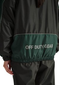 Person trägt eine schwarze und dunkelgrüne Jacke sowie Hose mit dem Aufdruck "OFF DUTY GEAR" auf der Rückseite in reflektierenden Buchstaben.