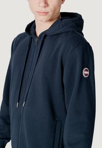 Felpa con zip color navy realizzata in tessuto morbido. Presenta un cappuccio con cordino, tasche laterali e una toppa circolare con il logo sulla manica sinistra.