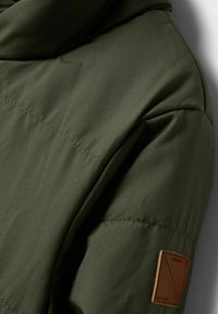 Nærbillede af ærmet på en olivengrøn polstret jakke med synlige sømme og en lille brun læderflapetiket.