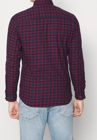 Uomo che indossa una camicia a maniche lunghe a quadri rossi e blu navy e jeans di un blu chiaro, mostrato di spalle su uno sfondo bianco semplice.