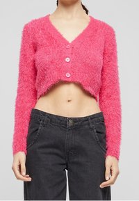 Kvinna som bär en fluffig rosa kortärmad cardigan med tre knappar och högmidjade svarta jeans, stående mot en neutral bakgrund.
