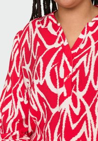 Röd blus med ett vitt abstrakt mönster, V-ringning och knappdetaljer på placket; tillverkad av lätt, texturerad tyg.