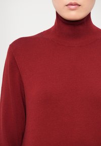 Piros garbó pulóver, puha kötött anyagból készült, testhezálló fazonú, hosszú ujjú és magas gallérral rendelkezik. Sima textúrájú, minták nélkül.