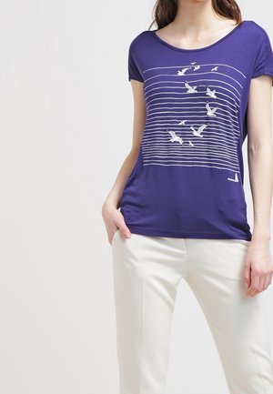 Vrouw die een paars shirt met korte mouwen draagt, met witte vogels en horizontale lijnen op de voorkant, gecombineerd met witte broek.