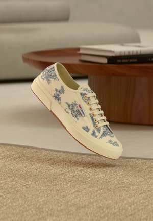 Sneaker in tela color crema con motivo toile de Jouy blu, lacci bianchi e suola in gomma che sembra fluttuare sopra un tappeto dalla trama evidente vicino a un tavolo di legno con libri.