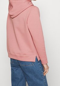 Sudadera con capucha rosa y un interior de forro polar texturizado. Presenta una abertura lateral y puños acanalados. Combinada con pantalones de mezclilla azules.