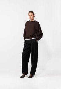 Kobieta stoi z rękami w kieszeniach, ubrana w ciemny, oversize'owy sweter, szerokie czarne spodnie, czarne eleganckie szpilki i wyraziste kolczyki na białym tle.