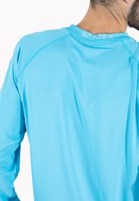 Lys blå langærmet træningsskjorte lavet af glat stof, med flade sømme og et lille "SPYDER" logo på bagsiden nær nakken.