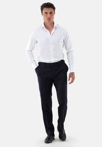 Camicia bianca a bottoni con maniche lunghe, abbinata a pantaloni blu navy. L'outfit è completato da scarpe eleganti nere. Design semplice e sartoriale.