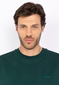 T-shirt di cotone verde scuro con collo rotondo e maniche corte, caratterizzato da un piccolo logo verde sul lato sinistro. Texture liscia.