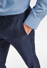 Pantalons taillés bleu marine en tissu texturé, avec poches latérales. Une chemise bleu clair à la finition lisse et avec des détails aux poignets est visible.