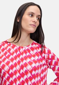 Blusa a maniche lunghe in tessuto leggero con un motivo geometrico rosa e rosso. Presenta un colletto rotondo e dettagli arricciati attorno alle spalle.