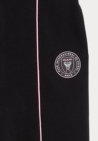 Pantalones cortos deportivos negros con una franja de acento rosa, que presentan un logo circular rosa con el texto "Miami" y el emblema del club. Textura de tela suave.