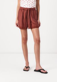 Pantaloni corti color ruggine con vestibilità ampia, cintura in tessuto e dettaglio ricamato all'orlo. Abbinati a ciabatte nere su uno sfondo semplice.