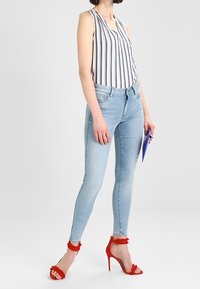 Jeans ajustados de color azul claro combinados con una blusa sin mangas a rayas. Presenta sandalias de tacón rojas con detalles de volantes. Sostiene una carpeta azul.