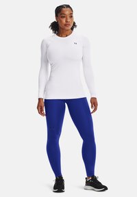 Langärmliges weißes Sportoberteil und blaue Leggings. Das Material wirkt glatt und enganliegend, mit unaufdringlicher Markenkennzeichnung auf beiden Teilen.