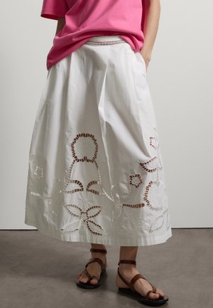 Person trägt einen weißen Midirock mit floralen Cut-Out-Mustern, ein teilweise in die Hose gestecktes rosa Hemd und braune Riemchensandalen.