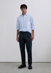 Cămașă albastru deschis cu nasturi și un logo mic, asortată cu pantaloni plisati verde închis și pantofi negri, stând pe un fundal neutru.