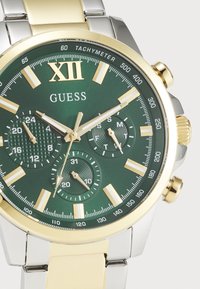 Gold and silver metal watch with a green dial. Features gold hour markers, sub-dials, tachymeter scale, and textured accents.