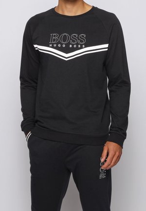 Uomo che indossa una felpa nera Hugo Boss con strisce a chevron bianche e pantaloni neri abbinati con il logo Hugo Boss sulla coscia.
