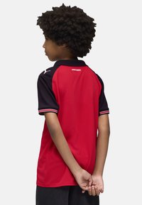 Camisa deportiva de manga corta en rojo y negro con cuello, con un tejido texturizado y ribetes blancos en las mangas. Logo en el hombro.