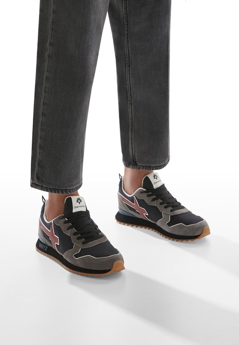 Personne portant des baskets noires et grises avec des étoiles bordeaux et des lacets noirs, associées à un pantalon gris foncé avec revers sur un sol blanc.