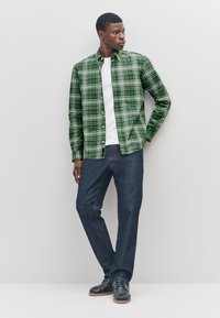 Green plaid shirt with white buttons, worn over a white T-shirt. Paired with blue denim jeans and black shoes with brown soles.