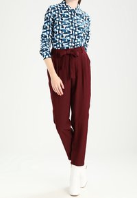 Long-sleeve shirt in blue with a geometric pattern, paired with high-waisted maroon trousers featuring a belt and soft texture. White boots.