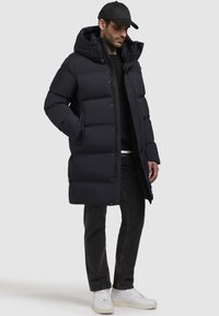 Cappotto imbottito blu navy con cappuccio, dotato di chiusura a zip e bottoni, tasche laterali e vestibilità comoda. Indossato con jeans scuri e sneakers bianche.